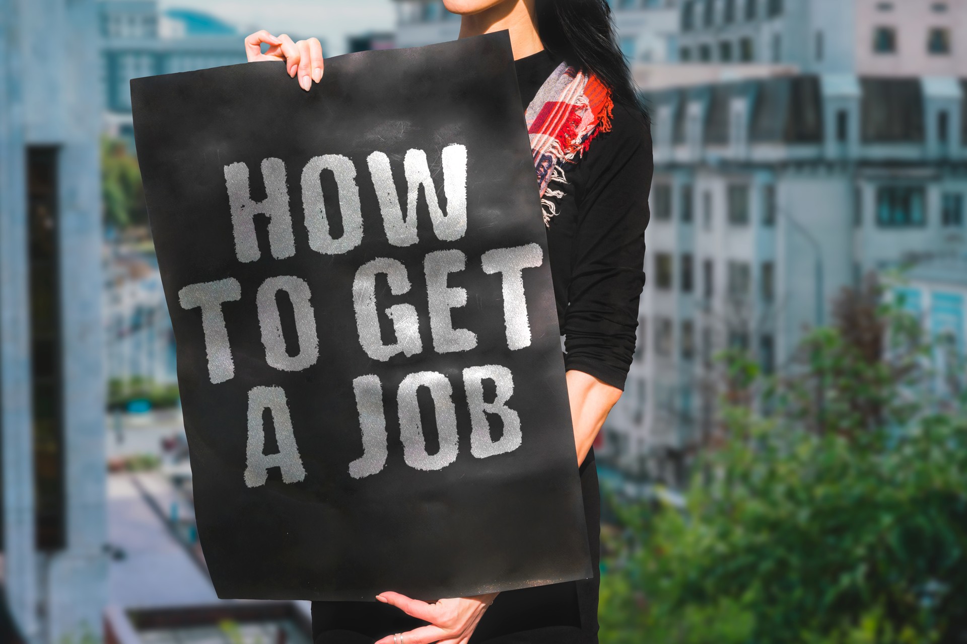 The phrase " How to get a job " is on a banner in women's hands. A woman holds black cardboard with an inscription. Problem. Office. Service. Find. Growth. Briefing. Help. Work. Leader. Mind. HR. Hire